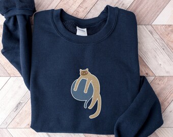 Op de afbeelding: Een donkerblauwe trui met een illustratie van een bruine kat die een blauwe bal vasthoudt. De kat kijkt naar rechts.