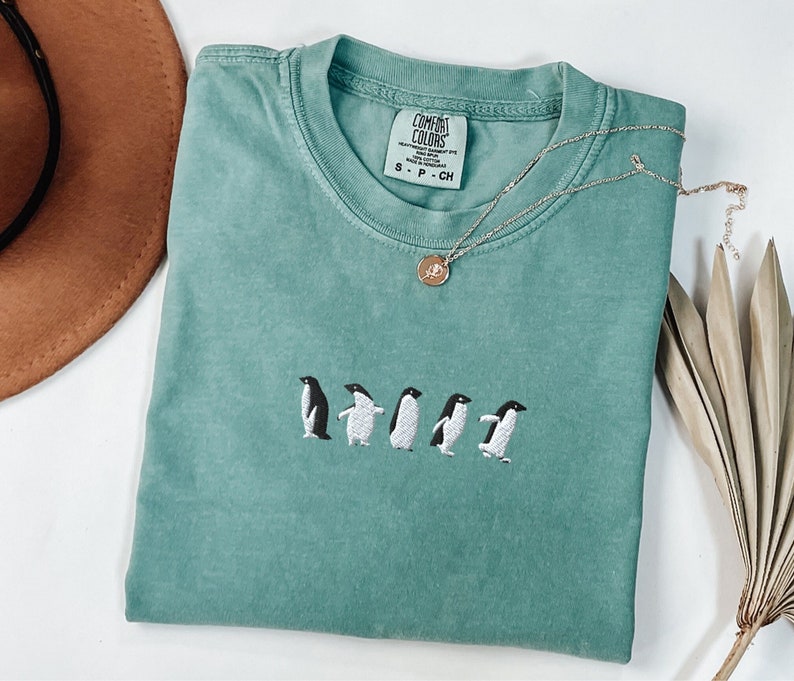 May include: A teal blue t-shirt with a line of five embroidered penguins in white. The shirt has the text "COMFORT COLORS" and "S - P - CH" on the tag.