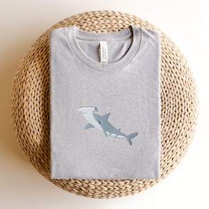 May include: A gray t-shirt with an embroidered hammerhead shark design. The shirt is folded and sitting on a woven straw mat.