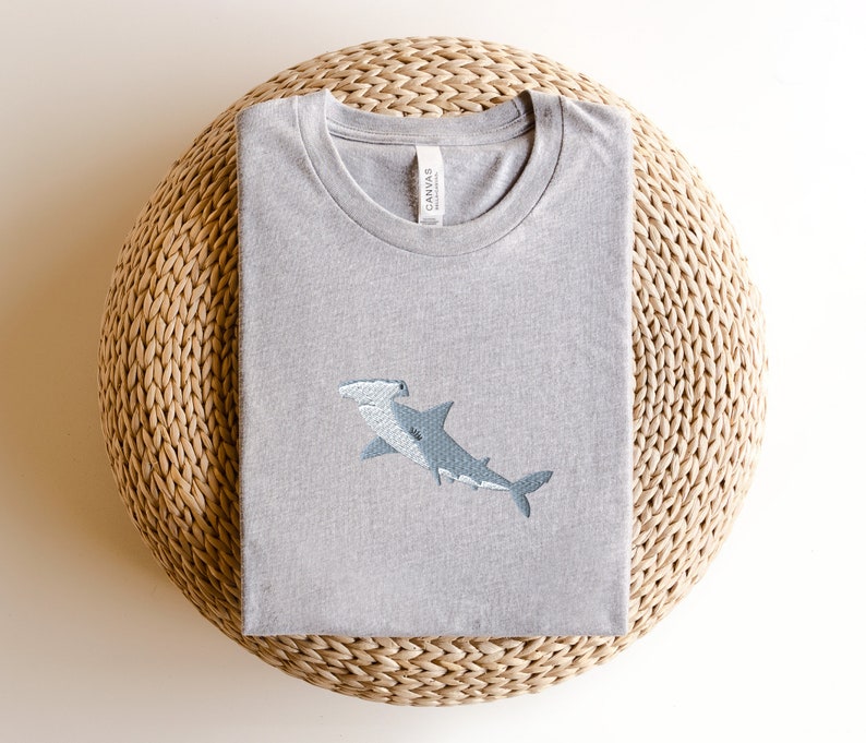 May include: A gray t-shirt with an embroidered hammerhead shark design. The shirt is folded and sitting on a woven straw mat.