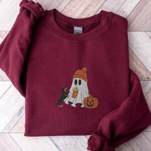May include: Burgundy sweatshirt with a white ghost wearing a red and orange hat holding a pumpkin and a black and brown dachshund looking at the ghost.
