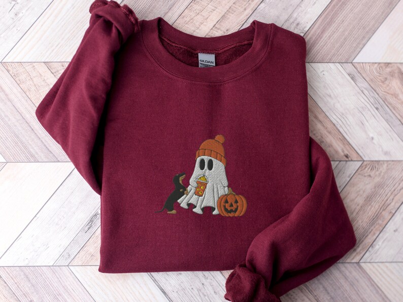 May include: Burgundy sweatshirt with a white ghost wearing a red and orange hat holding a pumpkin and a black and brown dachshund looking at the ghost.