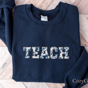 Op de afbeelding: Een marineblauw sweatshirt met het woord "TEACH" geborduurd op de voorkant in een wit en blauw bloemenpatroon. Het sweatshirt is opgevouwen op een lichtbruin en wit houten oppervlak.