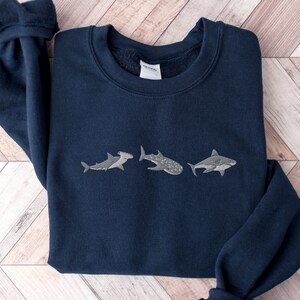May include: A navy blue sweatshirt with a white embroidered design of three different types of sharks. The sharks are a hammerhead, a whale shark, and a great white shark.