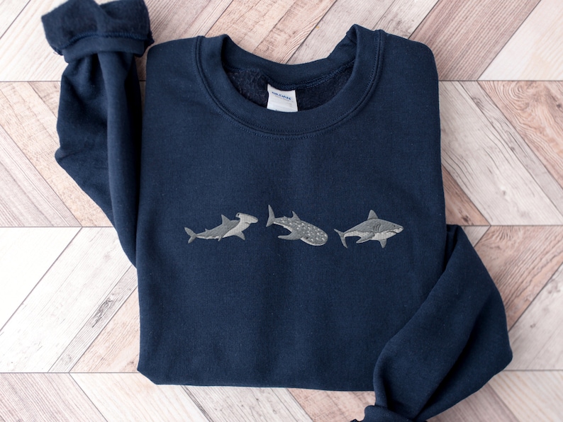 May include: A navy blue sweatshirt with a white embroidered design of three different types of sharks. The sharks are a hammerhead, a whale shark, and a great white shark.