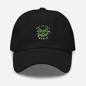 May include: A black baseball cap featuring an embroidered green cat face with a third eye on the forehead. The text "NOT OF THIS WORLD" is stitched in white in a circular pattern around the cat, accented by small white star and planet icons.