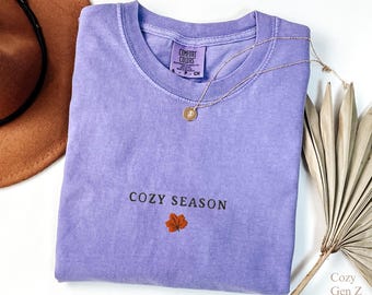 May include: A lavender Comfort Colors t-shirt with the words "COZY SEASON" and an embroidered orange leaf. A gold necklace and a brown hat are also in the image. The shirt is folded and ready to wear.