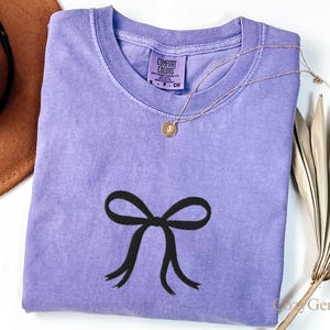 Könnte beinhalten: Ein lavendelfarbenes Comfort Colors T-Shirt mit einer schwarzen Schleife. Eine goldene Halskette mit Anhänger liegt auf dem Shirt. Ein brauner Hut und eine getrocknete Pflanze sind teilweise sichtbar. Das Etikett lautet "Comfort Colors".
