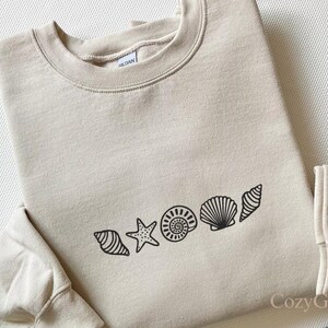 Könnte beinhalten: Beiges Sweatshirt mit Rundhalsausschnitt und schwarzem Muscheldesign. Das Design umfasst einen Seestern, eine Spiralmuschel und andere Muschelformen. Der Markenname "Gildan" ist auf dem Etikett sichtbar. Das Sweatshirt ist aus weichem Material.