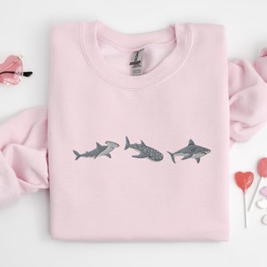 May include: Pink sweatshirt with a graphic of three sharks embroidered on the front. The sharks are a hammerhead, a whale shark, and a great white shark.