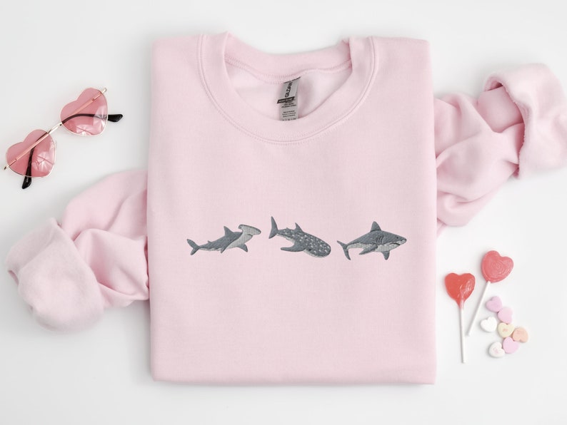 May include: Pink sweatshirt with a graphic of three sharks embroidered on the front. The sharks are a hammerhead, a whale shark, and a great white shark.