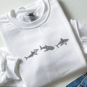 May include: White sweatshirt featuring a graphic of four different sharks in a line. The sharks are grey and white and are facing the right side of the image.