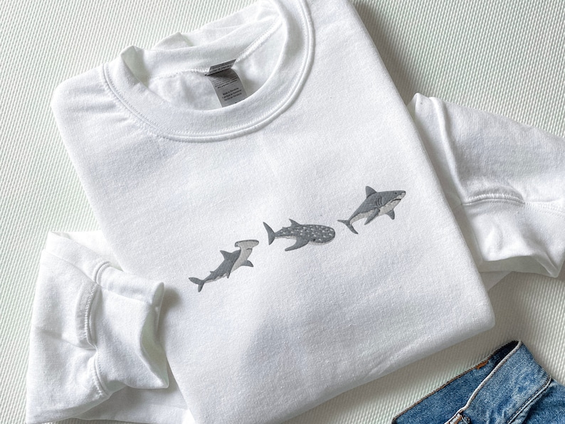 May include: White sweatshirt featuring a graphic of four different sharks in a line. The sharks are grey and white and are facing the right side of the image.