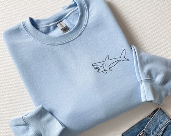 Könnte beinhalten: Ein hellblauer Pullover mit einer einfachen schwarzen Linienzeichnung eines Hais auf der Vorderseite.