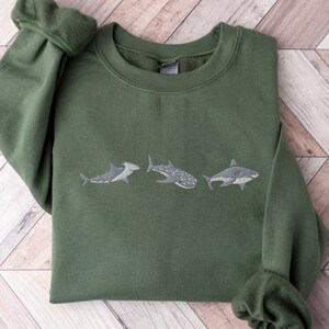 May include: A green sweatshirt featuring an embroidered design of three different types of sharks. The sharks are arranged in a row, with the hammerhead shark on the left, the whale shark in the middle, and the great white shark on the right.