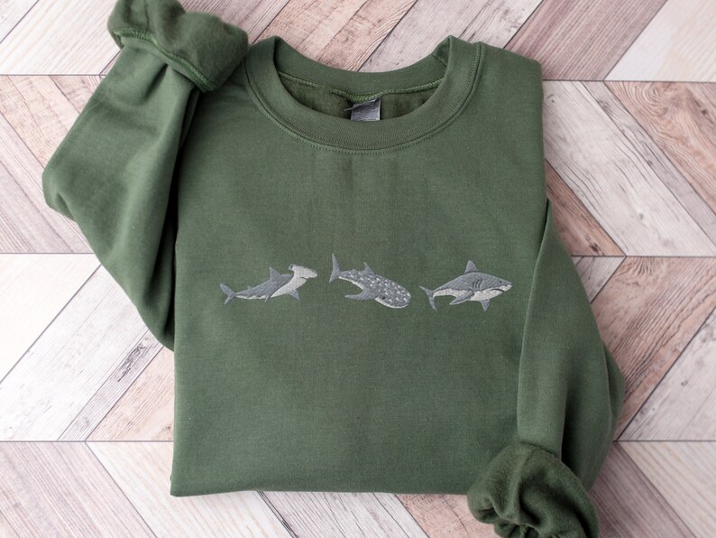 May include: A green sweatshirt featuring an embroidered design of three different types of sharks. The sharks are arranged in a row, with the hammerhead shark on the left, the whale shark in the middle, and the great white shark on the right.