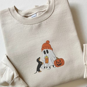 May include: A beige sweatshirt with a graphic of a ghost wearing a red hat and holding a cup, a black dachshund looking at the ghost, and a jack-o-lantern.