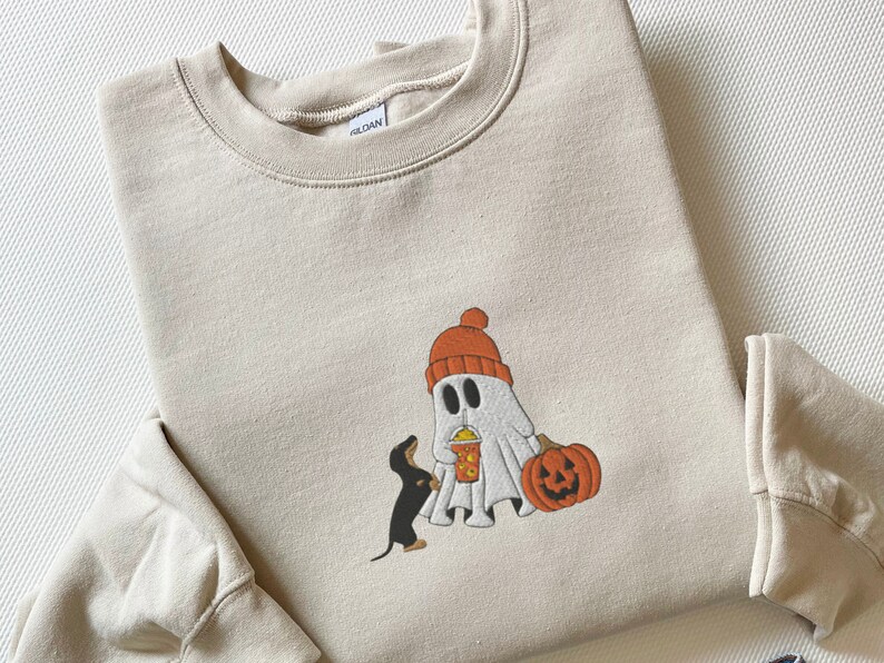 May include: A beige sweatshirt with a graphic of a ghost wearing a red hat and holding a cup, a black dachshund looking at the ghost, and a jack-o-lantern.