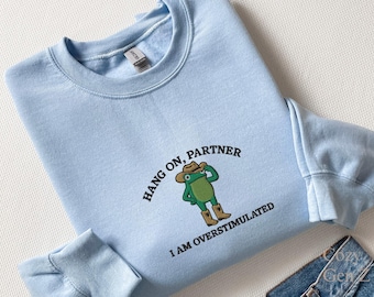 May include: Light blue sweatshirt with the text "Hang on, Partner, I am overstimulated." The design features a cartoon frog wearing a cowboy hat and boots. The sweatshirt is made of a soft material.