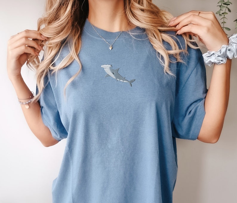 May include: A light blue t-shirt with an embroidered hammerhead shark design. The shirt has a relaxed fit and short sleeves.