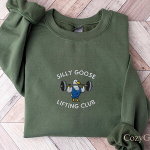 Könnte beinhalten: Olivgrünes Sweatshirt mit dem gestickten Text "SILLY GOOSE LIFTING CLUB". Das Design zeigt eine Cartoon-Gans, die Gewichte hebt. Das Sweatshirt ist auf einer Holzoberfläche gefaltet.