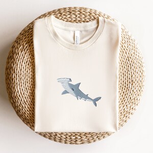May include: A white t-shirt with a blue hammerhead shark embroidered on the front. The shirt is on a woven straw mat.
