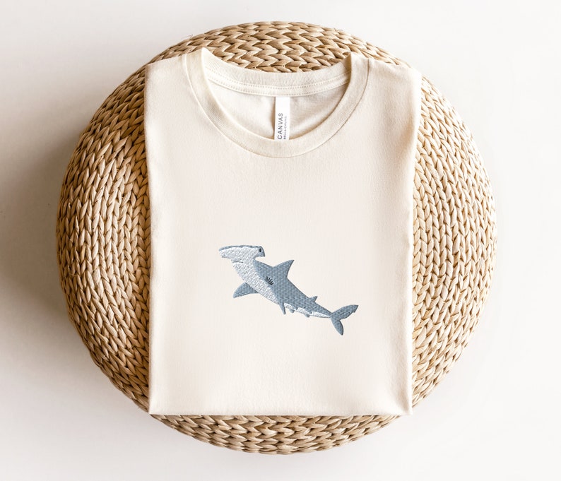 May include: A white t-shirt with a blue hammerhead shark embroidered on the front. The shirt is on a woven straw mat.