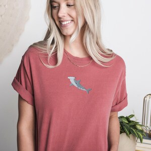 May include: A red t-shirt with a hammerhead shark embroidered on the front. The shark is gray and white.