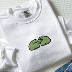 May include: White sweatshirt with a green embroidered design of two frogs hugging.