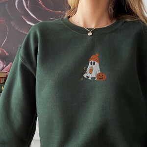 May include: A dark green sweatshirt with a white ghost wearing a red and white hat holding a pumpkin. The ghost is embroidered on the front of the sweatshirt.