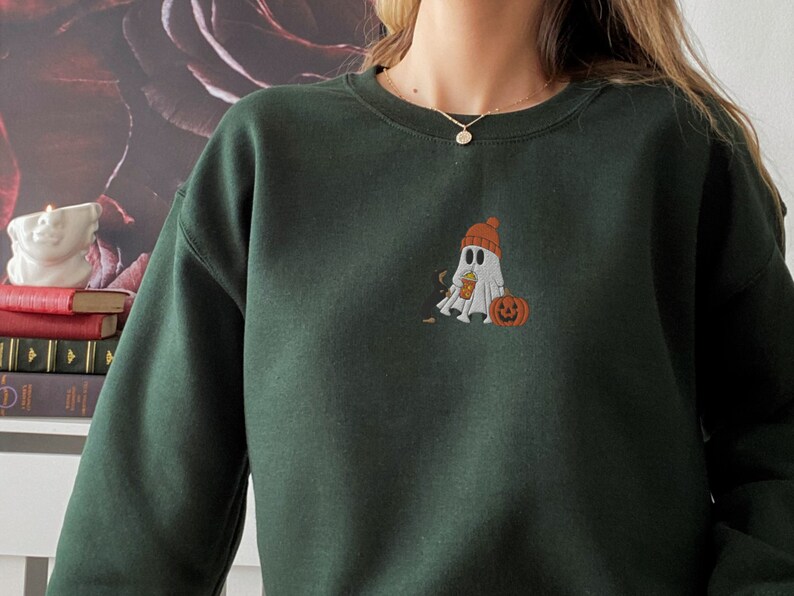 May include: A dark green sweatshirt with a white ghost wearing a red and white hat holding a pumpkin. The ghost is embroidered on the front of the sweatshirt.