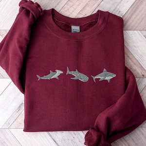 May include: Burgundy sweatshirt with three embroidered sharks: a hammerhead, a whale shark, and a great white shark.