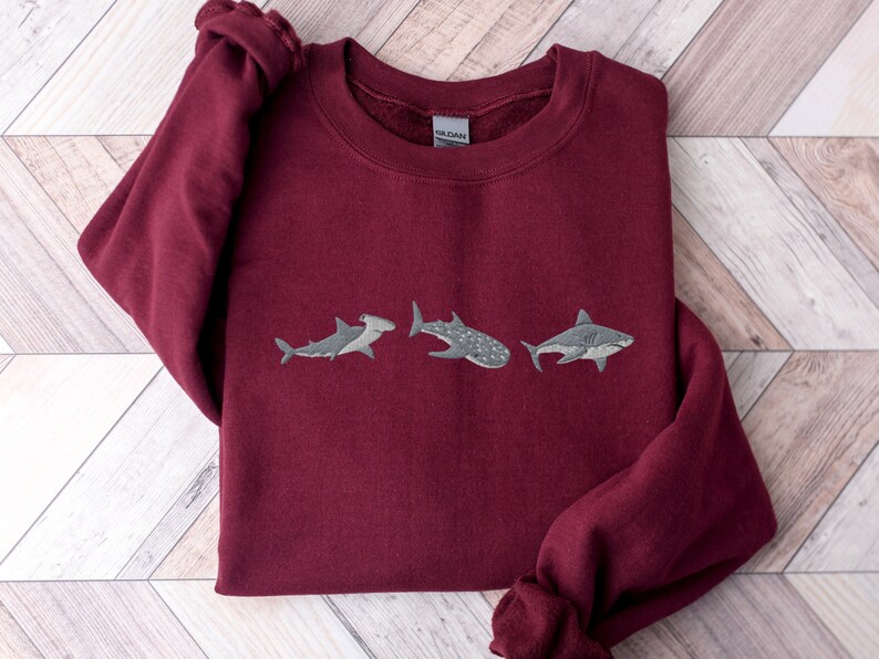 May include: Burgundy sweatshirt with three embroidered sharks: a hammerhead, a whale shark, and a great white shark.