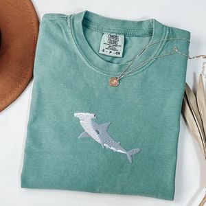 May include: A teal blue t-shirt with a white embroidered hammerhead shark. The shirt has a tag that reads "COMFORT COLORS" and "S - P - CH".