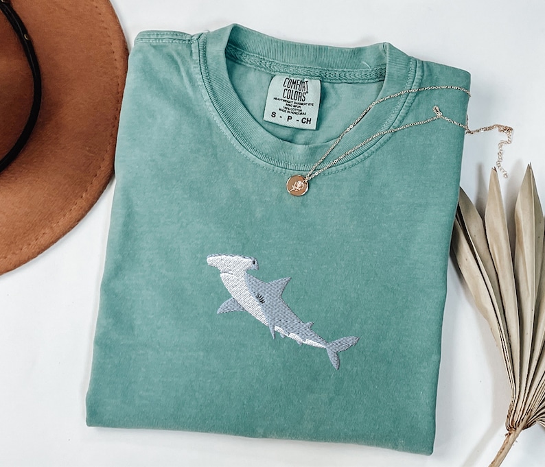 May include: A teal blue t-shirt with a white embroidered hammerhead shark. The shirt has a tag that reads "COMFORT COLORS" and "S - P - CH".
