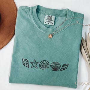 Könnte beinhalten: Ein türkisgrünes T-Shirt mit einem schwarzen Muschelgrafik. Das Shirt hat einen Rundhalsausschnitt und kurze Ärmel. Eine goldene Halskette mit Anhänger liegt über dem Shirt. Das Shirt ist auf einer weißen Oberfläche gefaltet.