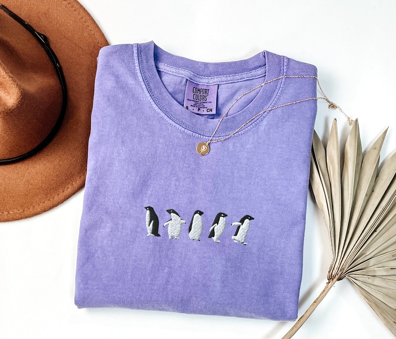 May include: A lavender t-shirt with a line of five embroidered penguins. The shirt has a tag that reads "Comfort Colors" and a silver necklace with a circular pendant.