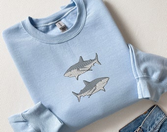 Pode incluir: Uma camisa de moletom azul claro com dois tubarões cinzentos bordados na frente.