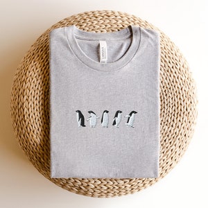 May include: A heather grey t-shirt with an embroidered design of five penguins walking in a line. The t-shirt has a crew neck and is made by Canvas.