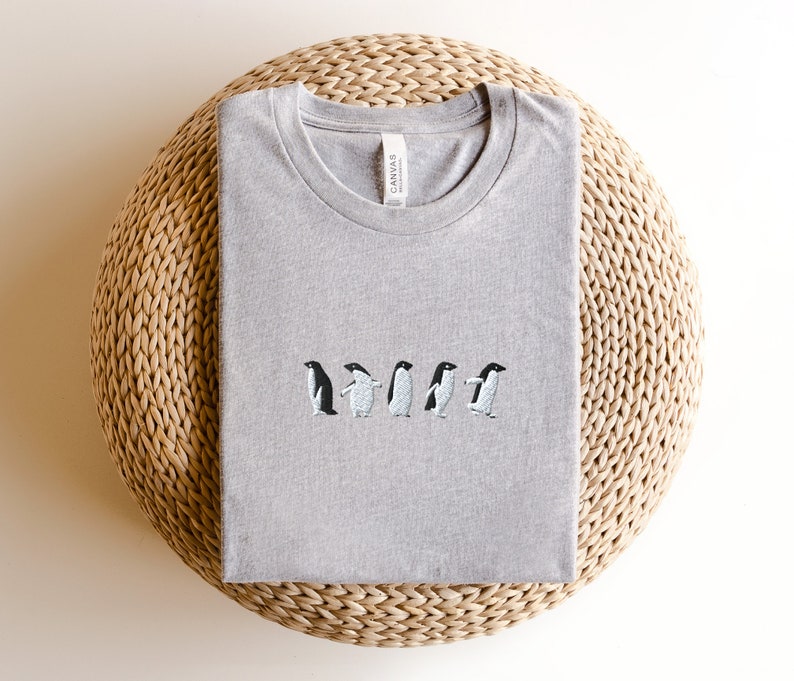 May include: A heather grey t-shirt with an embroidered design of five penguins walking in a line. The t-shirt has a crew neck and is made by Canvas.
