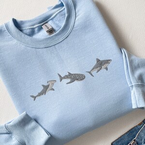 May include: A light blue sweatshirt featuring a graphic of three different types of sharks: a hammerhead, a whale shark, and a great white shark.
