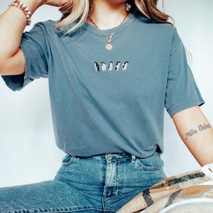 May include: A woman wearing a blue t-shirt with a line of embroidered penguins. She is wearing blue jeans and a gold necklace with a round pendant. The woman has a tattoo on her arm that says "Mon Ocean".