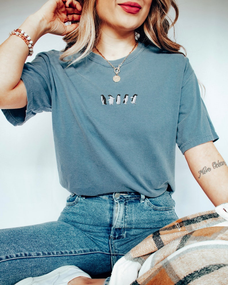 May include: A woman wearing a blue t-shirt with a line of embroidered penguins. She is wearing blue jeans and a gold necklace with a round pendant. The woman has a tattoo on her arm that says "Mon Ocean".