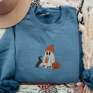 May include: A blue sweatshirt with a ghost and a dog wearing a hat and holding a pumpkin. The ghost is holding a cup of popcorn.