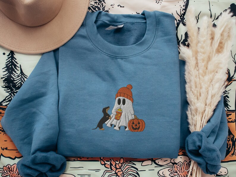 May include: A blue sweatshirt with a ghost and a dog wearing a hat and holding a pumpkin. The ghost is holding a cup of popcorn.