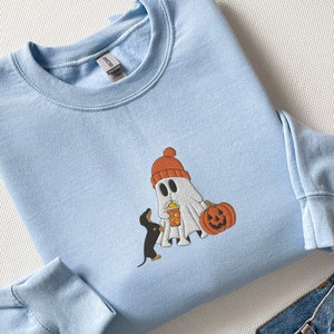 May include: A light blue sweatshirt with a cartoon ghost wearing a red hat and holding a pumpkin and a cup. A small black and brown dachshund is standing next to the ghost.