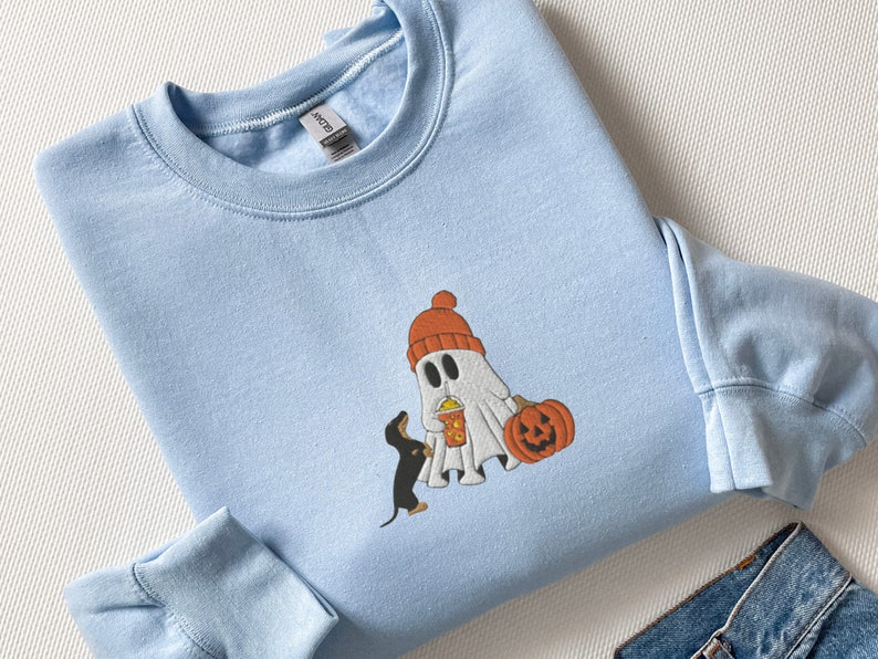 May include: A light blue sweatshirt with a cartoon ghost wearing a red hat and holding a pumpkin and a cup. A small black and brown dachshund is standing next to the ghost.