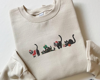 May include: A beige sweatshirt with a fun design of black cats in Christmas outfits. The cats wear Santa hats, red coats, and are surrounded by Christmas presents, creating a festive holiday theme. The sweatshirt is made of a soft material.