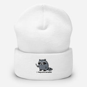 May include: White beanie featuring an embroidered cartoon raccoon holding a knife. The raccoon is gray and black with a white belly. Below the raccoon is the text "I require snacks".