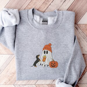 May include: A gray sweatshirt with a cute ghost wearing a red hat and holding a cup of popcorn. A small black dog is standing next to the ghost. A pumpkin is in front of the ghost.